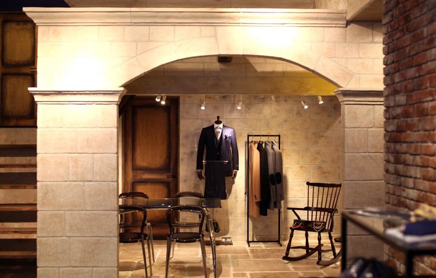アーチ状の壁と石調の内装が印象的な店舗空間。スーツのトルソーやハンガーラック、テーブルと椅子を配置した落ち着いた雰囲気の内装デザイン。