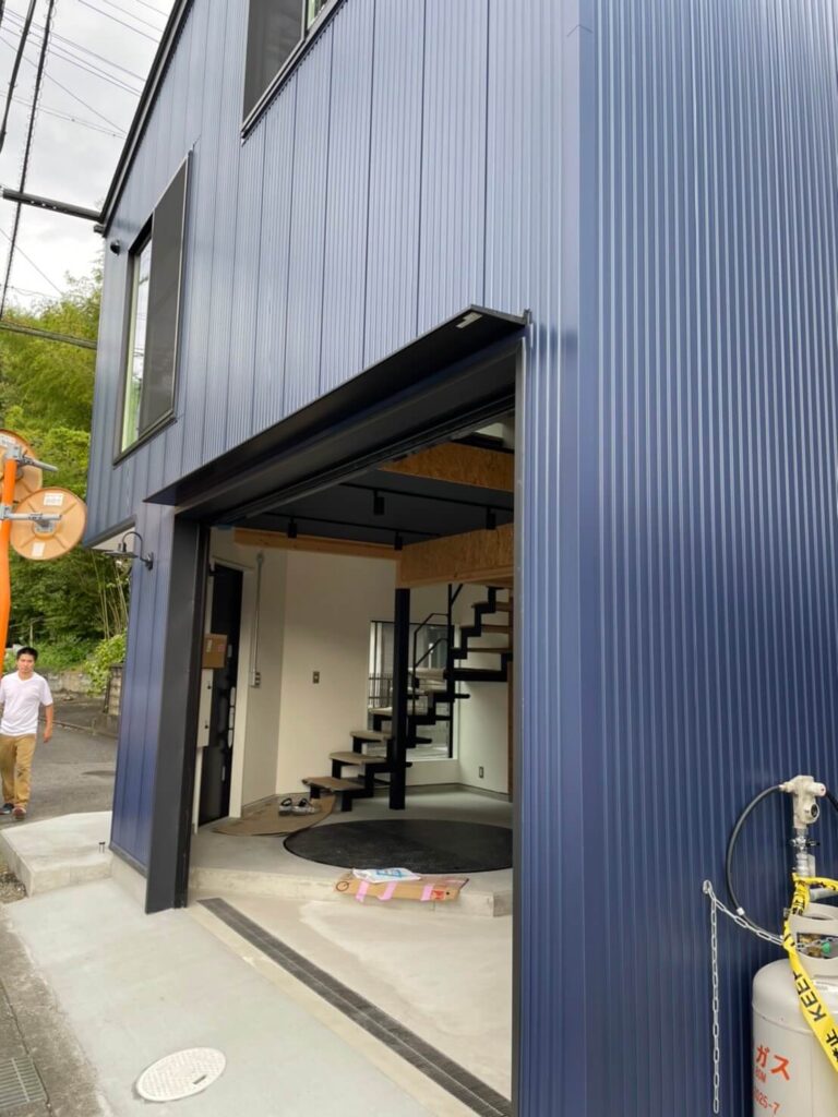 縦リブのネイビーカラー外壁を持つガルバリウム鋼板の建物外観。大開口のガレージシャッターが開放され、内部には黒い鉄骨のスケルトン階段と吹き抜け空間、OSB合板のロフト手すりが見える。土間コンクリート床とエントランス前の排水溝、右側に設置されたプロパンガスボンベ、左奥の道路と人物が写っている施工直後の様子。