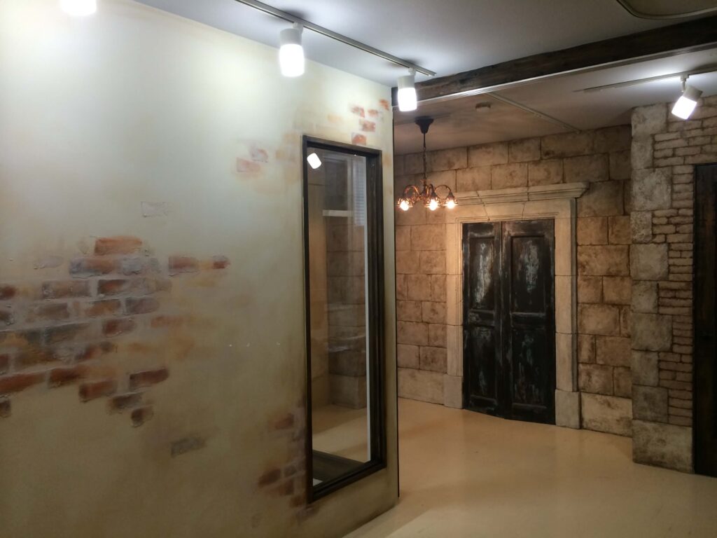 エイジング塗装の壁と石積み風パネルで構成されたスタジオ風の内装空間。黒枠の室内窓、アンティーク調のシャンデリア照明、重厚感のあるダブルドアが配置され、天井のスポットライトが質感を強調するヴィンテージテイストのデザイン。