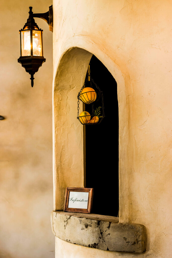 塗り壁のアーチ型ニッチに、チェーンで吊られたワイヤーバスケットと果物のディスプレイ、下部のモルタル棚に小さなフレームが置かれた装飾。左側にはアンティーク調の壁付けランタン照明が灯り、温かみのある南欧風の内装デザインを演出している。