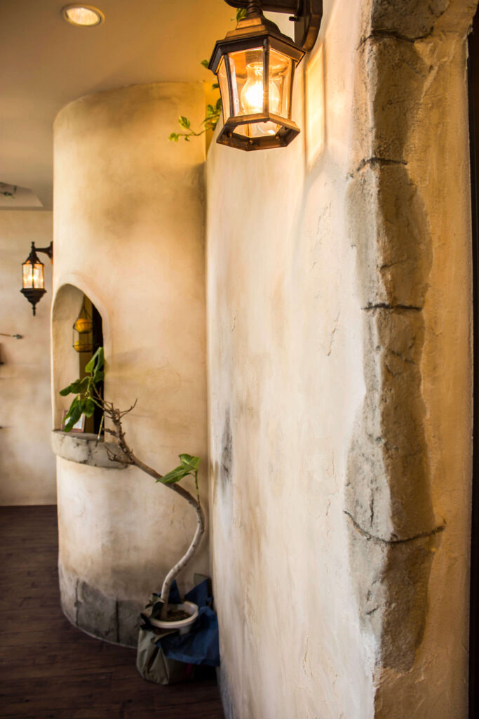 エイジング仕上げの塗り壁と石積み風の角柱が続く内装通路。壁付けのアンティーク調ランタン照明が温かく灯り、アーチ型ニッチや観葉植物が配置された南欧テイストの落ち着いた空間デザイン。