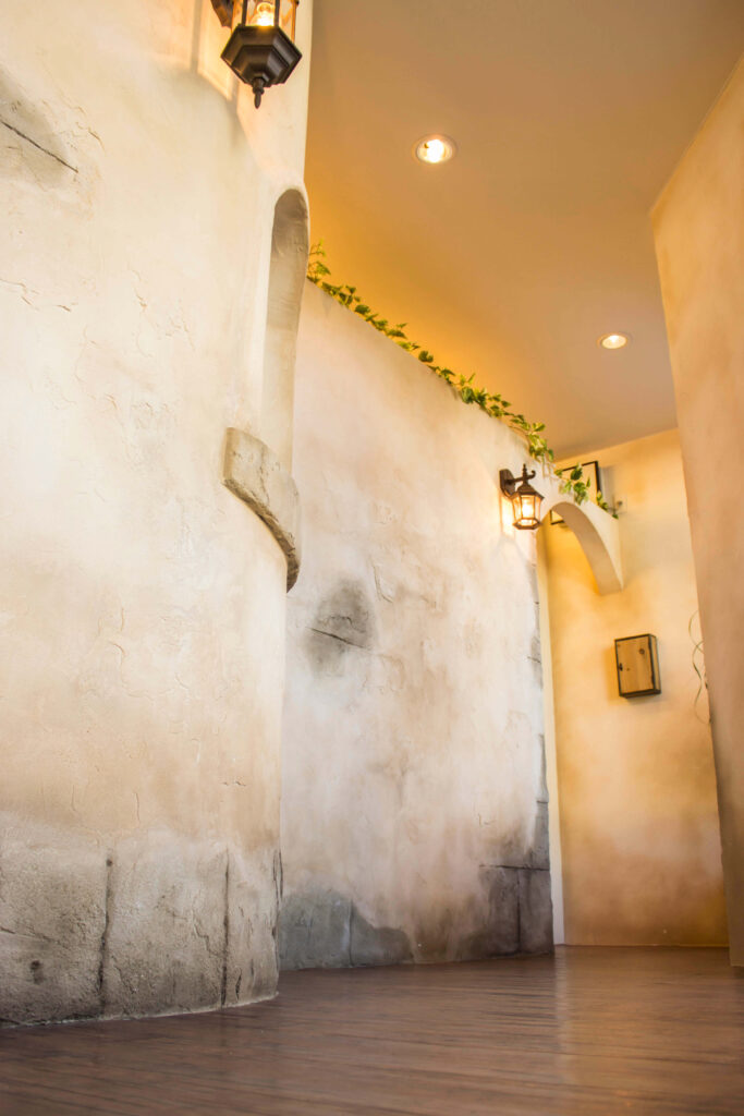 エイジング塗装の白い塗り壁が続く通路空間。壁付けのアンティーク調ランタン照明、上部のグリーン装飾、アーチ開口が奥へと連なり、ダークブラウンの床材と暖色ダウンライトが南欧風の雰囲気を演出している。