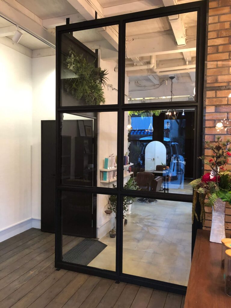 黒いスチールフレームのガラスパーテーション越しに見える美容室内装。白い天井配管と観葉植物、スタイリングチェアやカウンターが配置され、外のウッドデッキ床とレンガ壁がインダストリアルな雰囲気を演出している空間。