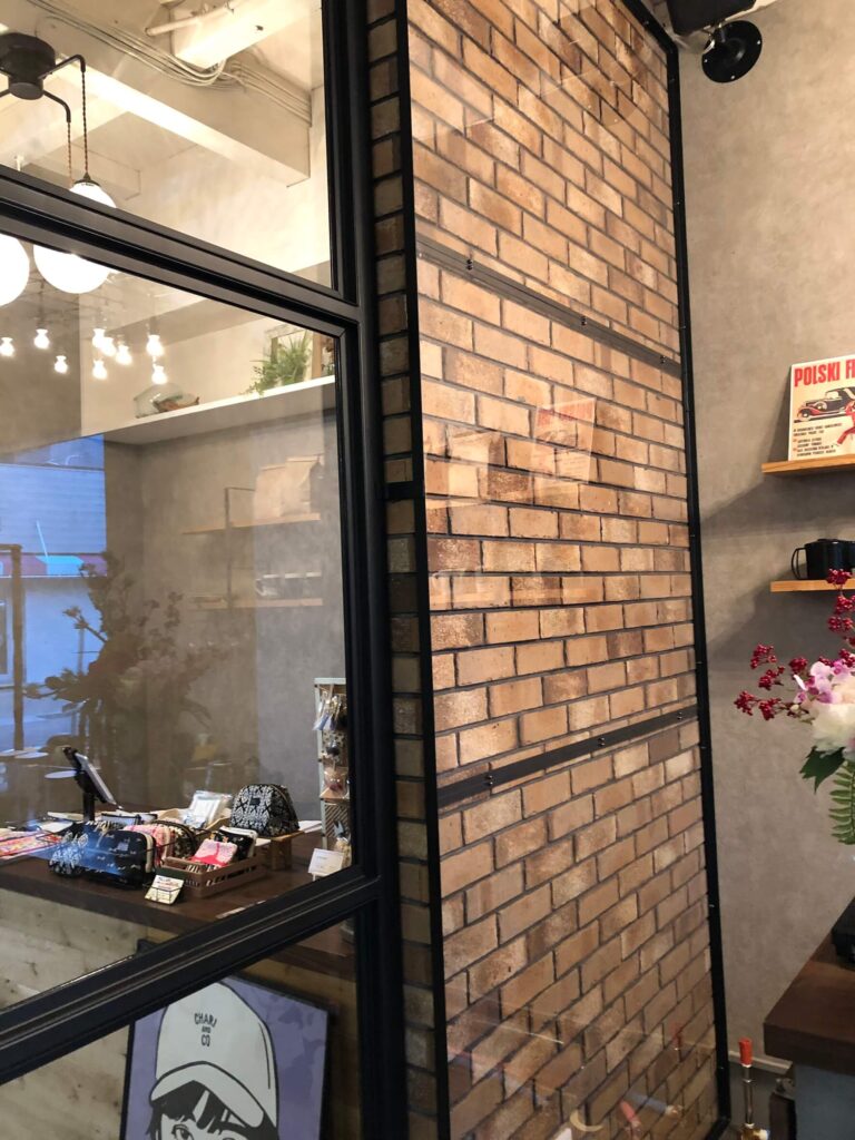 黒いスチールフレームのガラスとレンガ壁が組み合わさったインダストリアルな内装。ガラス越しに雑貨が並ぶカウンターや棚が見え、モルタル調の壁とスポット照明が落ち着いたショップ空間を演出している。
