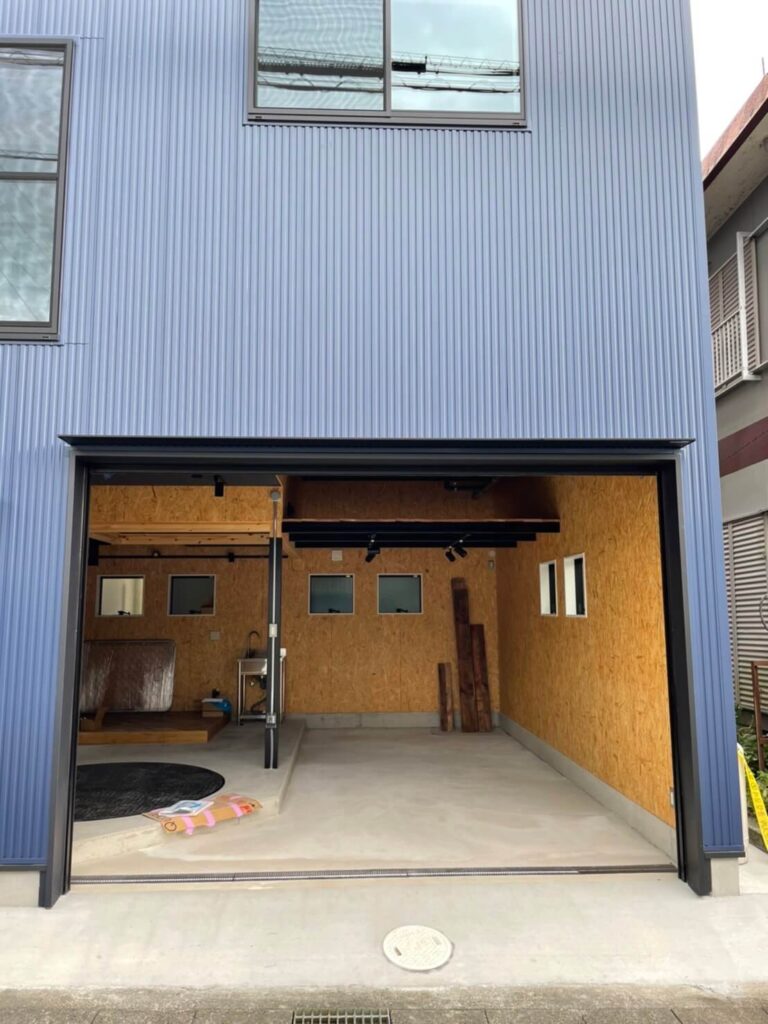 縦リブのネイビーカラー外壁を持つガルバリウム鋼板の建物外観。大開口のガレージシャッターが開放され、内部には黒い鉄骨のスケルトン階段と吹き抜け空間、OSB合板のロフト手すりが見える。土間コンクリート床とエントランス前の排水溝、右側に設置されたプロパンガスボンベ、左奥の道路と人物が写っている施工直後の様子。