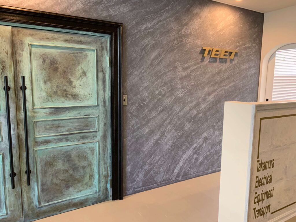 エイジング加工の重厚な両開きドアと、石目調のグレーアクセントウォールが印象的なエントランス内装。壁面にはロゴサイン、右側にはクラシックな装飾枠の受付カウンターがあり、上品で落ち着いた雰囲気を演出している空間。