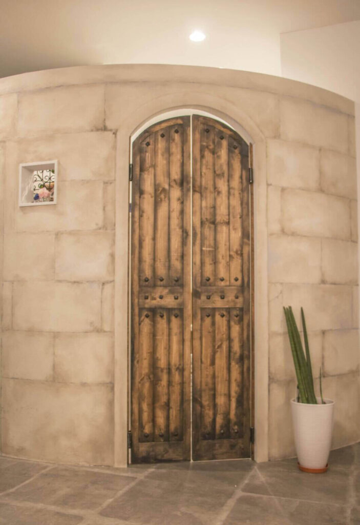 石積み風のアーチ壁に収まる、アイアンスタッズ付きの両開き木製ドア。床は石調タイル、右側に白い鉢植えの観葉植物が置かれ、南欧テイストの重厚感ある内装エントランス。