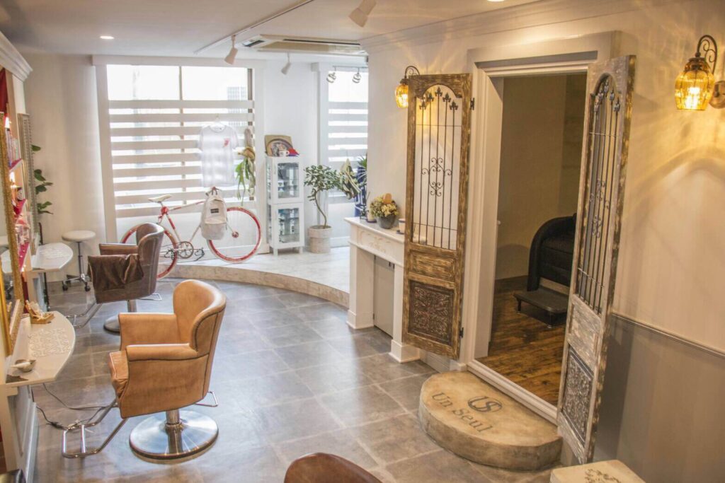 南欧テイストの美容室内装。アイアン装飾付きのアンティークドアが開いた個室入口、石調タイル床と曲線ステップ、ブラウンのスタイリングチェア、奥の大きな窓際には自転車や観葉植物、ディスプレイ棚が配置された明るく温かみのある空間。