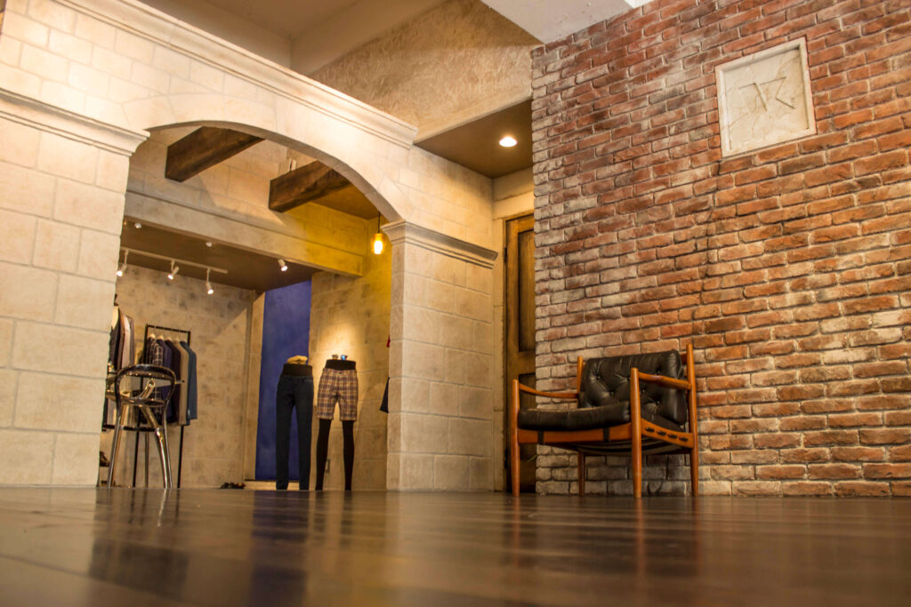 レンガ調のアクセントウォールと石目調のアーチ開口が特徴的な店舗内装。ダークブラウンのフローリング床、ヴィンテージ風のレザーラウンジチェア、奥にはアパレル什器とマネキンに着せられた衣類、天井のスポットライトと温かみのあるペンダント照明が空間を照らすクラシックモダンなデザイン。