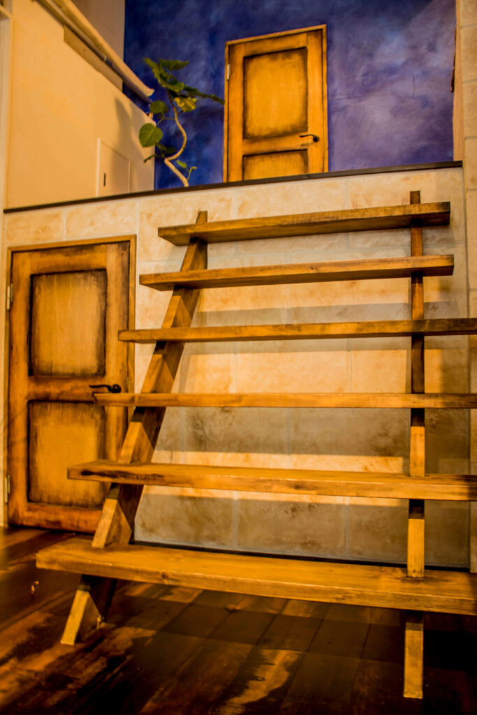 モルタル調の壁面に造作された木製の段違いディスプレイ棚と、エイジング加工の木製ドアが並ぶ店舗内装。上部ロフトには深いブルーの塗り壁と観葉植物、アンティーク調のドアが見え、ダークウッドの床材と温かみのある照明がヴィンテージ感を演出している。