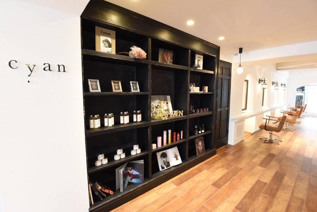 白い塗り壁と温かみのある木目フローリングの美容室内装。壁一面に設置されたブラックの造作シェルフにはヘアケア商品や写真、装飾品が並び、右側にはアーチ開口とブラケット照明が続くカットスペースにブラウンのスタイリングチェアが整列している明るく上品な空間。