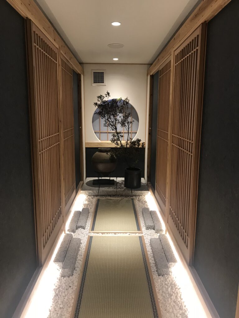 木格子の建具が並ぶ廊下の中央に畳敷きと白砂利を配し、奥の丸窓と植栽が見える和モダン空間。足元の間接照明が通路をやわらかく照らしている。