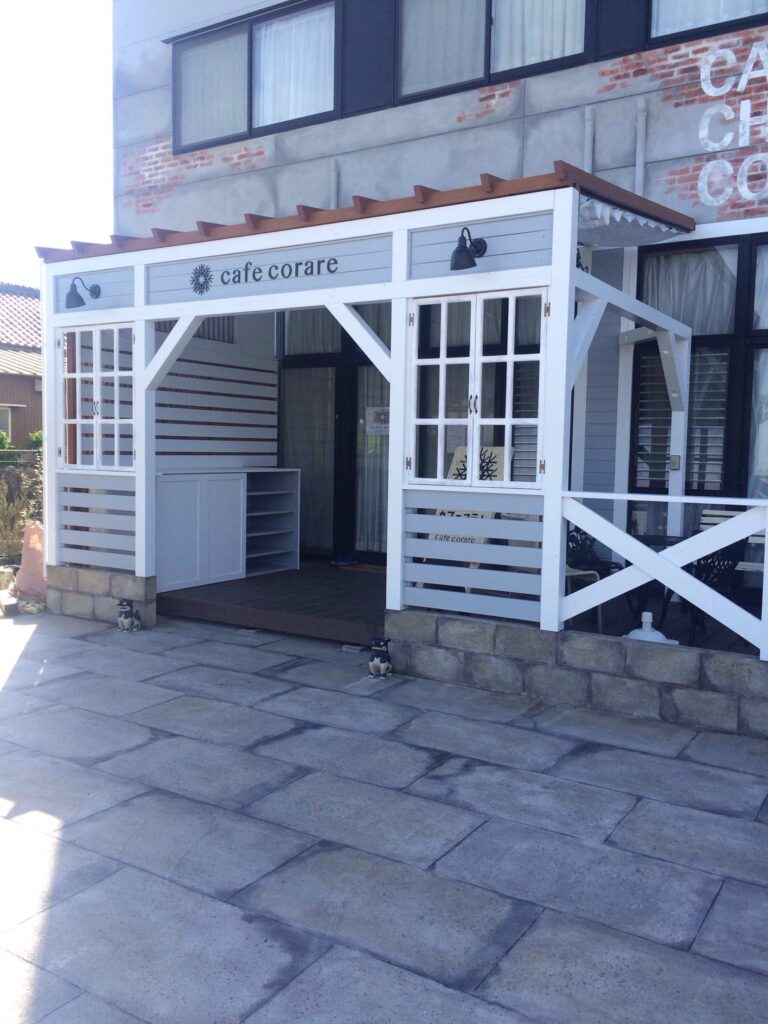 白い木製フレームとグレーの板張りで造作された『cafe corare』のテラス風ファサード。格子窓やブラケット照明が付いたナチュラルな店舗外観。