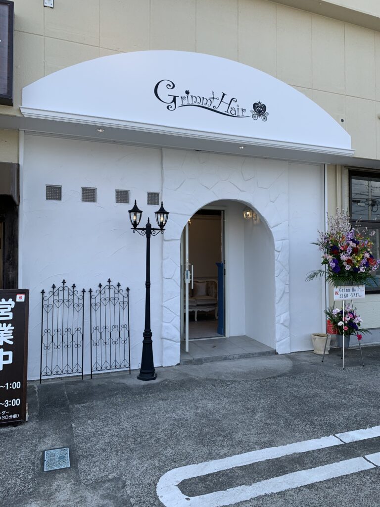 白い塗り壁とアーチ入口、アイアン装飾が特徴の『Grimm Hair』店舗外観。クラシックな街灯風照明と花スタンドが並ぶ可愛らしいファサード。