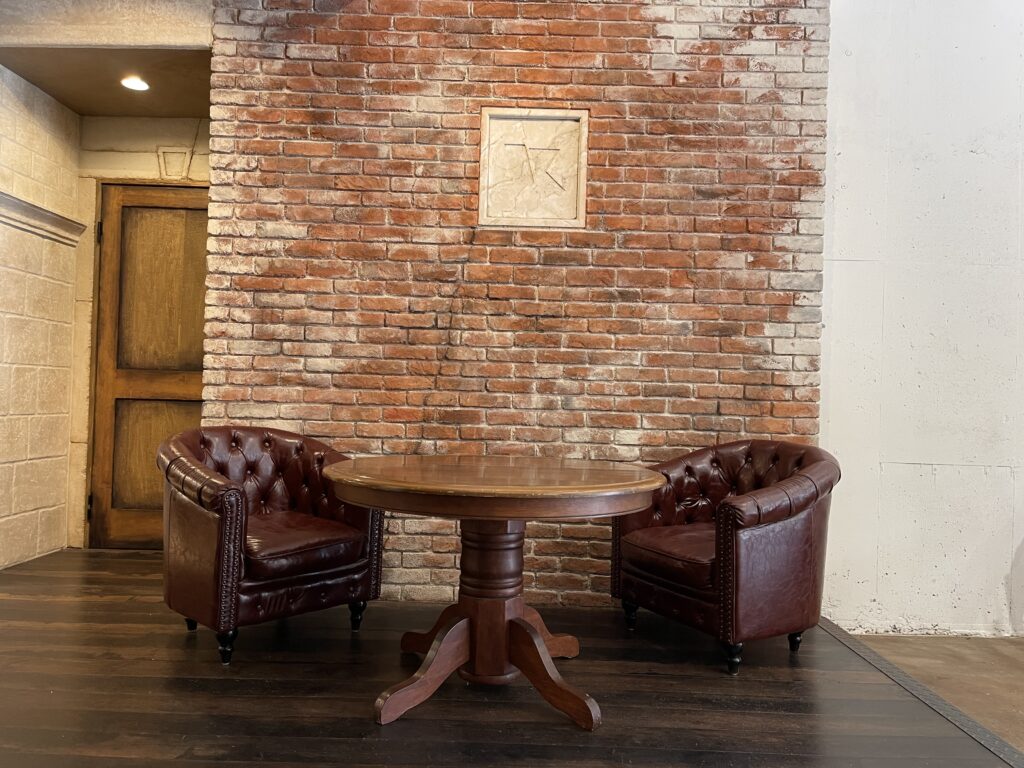 レンガ壁を背景に、丸い木製テーブルとブラウンのレザーチェアが向かい合う落ち着いたヴィンテージ風の打ち合わせスペース。