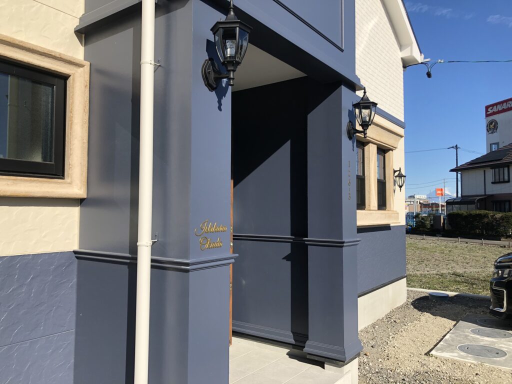 ネイビーの外壁とベージュのモールディングが印象的な洋風住宅の外観。クラシックな壁付けランタン照明と金色の表札が設置された玄関ポーチ、石張り風の窓枠がアクセントになっている。