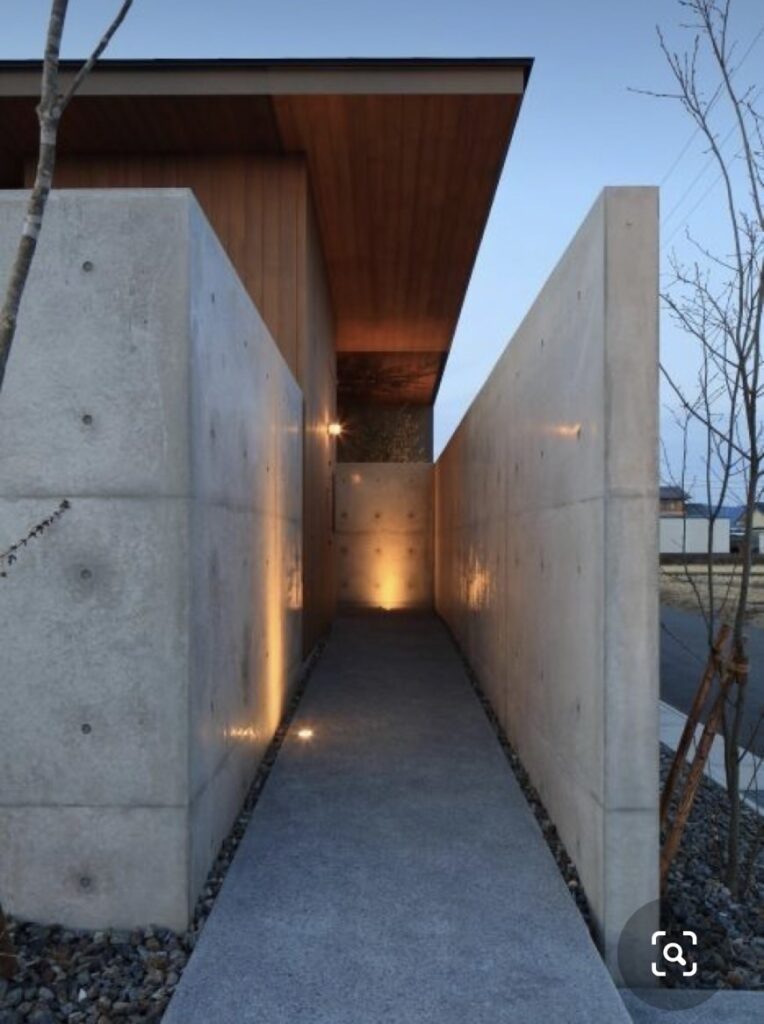 打ち放しコンクリートの壁に囲まれた細長いアプローチ通路。足元と壁面の間接照明が夕暮れに柔らかく灯り、木張りの軒天が奥行きを強調するミニマルモダンな外構デザイン。