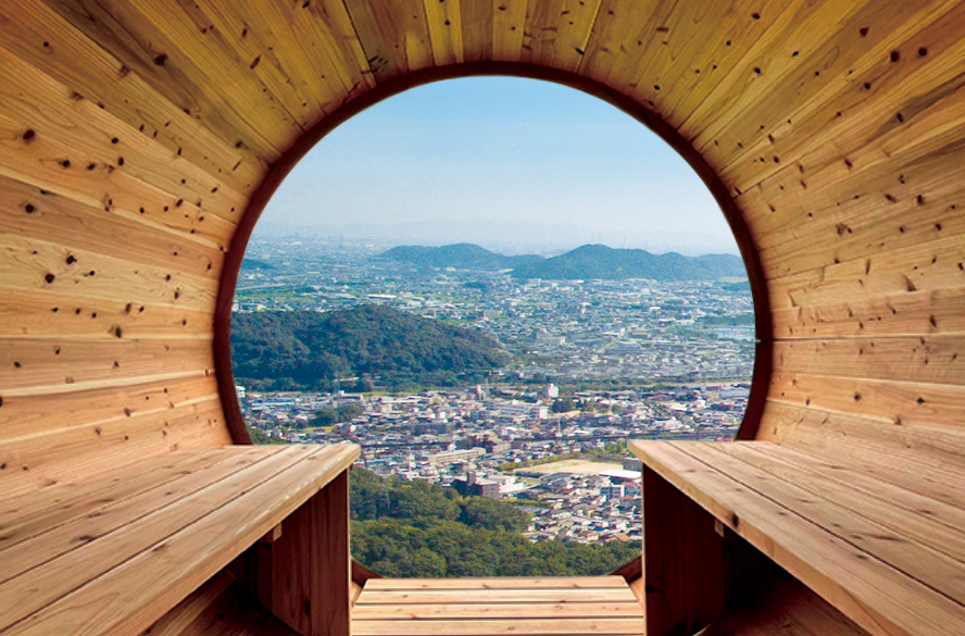 木板張りのトンネル状展望スペースの丸い開口から、山々と市街地を一望する景色。左右に木製ベンチが設けられ、自然素材に包まれたフレーミングが眺望を強調している。