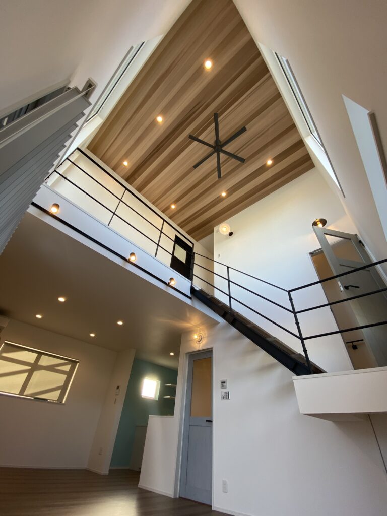 吹き抜けのあるモダンなリビング内装。木目の勾配天井にシーリングファンとダウンライト、黒いアイアン手すりの階段と2階廊下、白壁と木床が開放感を演出している空間。