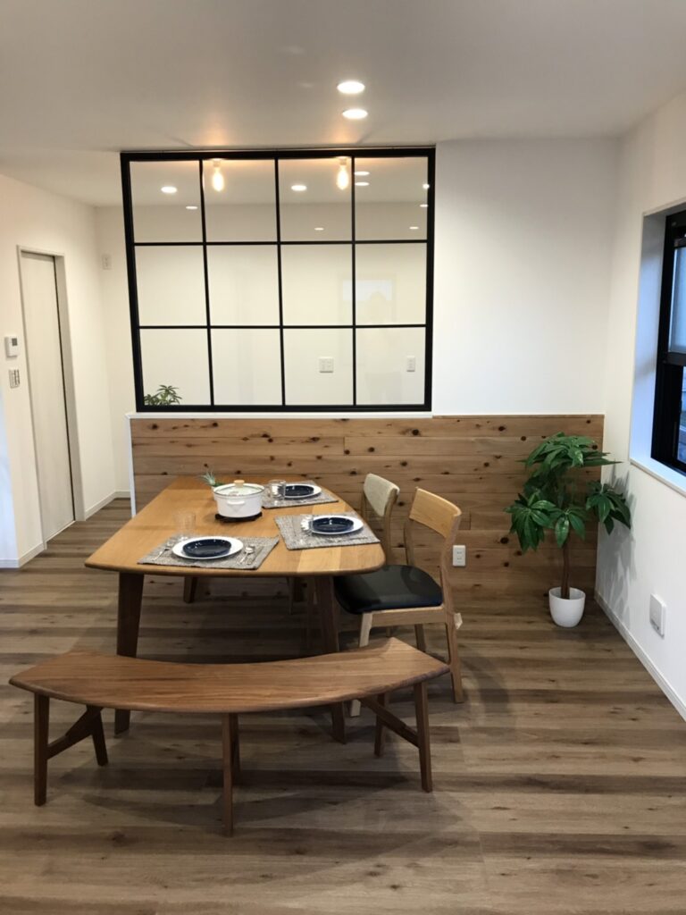 木目フローリングと腰板、黒い格子の室内窓がアクセントのダイニング空間。無垢材テーブルとベンチ、観葉植物が配置され、白壁とダウンライトで温かみのあるナチュラルモダンな内装。