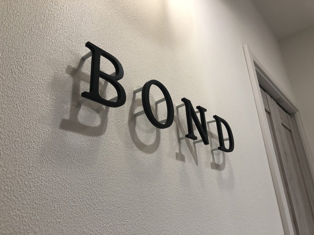白い凹凸のある壁面に、立体的な黒い『BOND』の文字サインが浮かせて取り付けられ、照明で影が美しく落ちている内装ディテール。