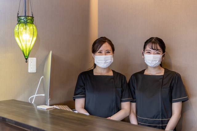 受付カウンターに並んで立つマスク着用の女性スタッフ2名が迎えるサロンの受付風景