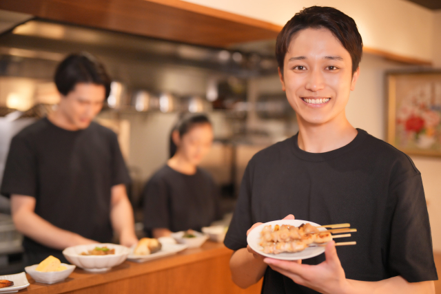 温かみのあるオープンキッチンで、焼き鳥を手に笑顔で提供するスタッフと、奥で調理をするチームの様子