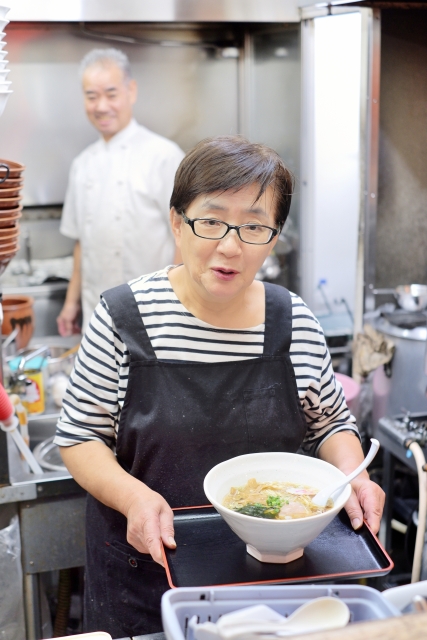 厨房で出来上がったラーメンをお盆にのせて提供する女性店主と、奥で調理を続ける男性スタッフの様子
