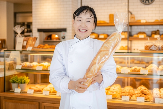 焼きたてパンが並ぶ店内で、大きなフランスパンを手に笑顔を見せる女性パン職人