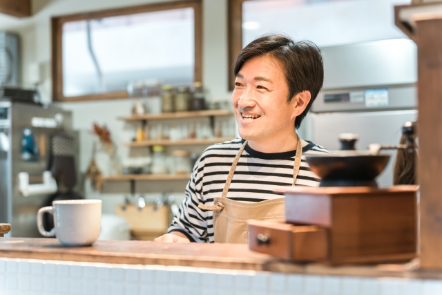 カフェカウンターで笑顔で接客する男性スタッフ。木製のコーヒーミルとマグカップが置かれた温かみのある店内風景
