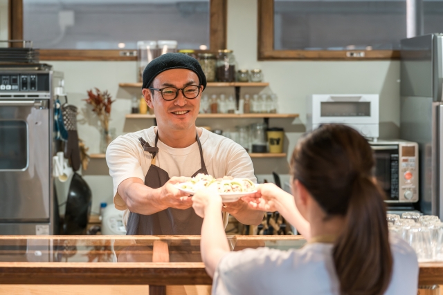 カウンター越しに料理を手渡す店主と受け取るお客様の様子が写る温かみのある飲食店のシーン